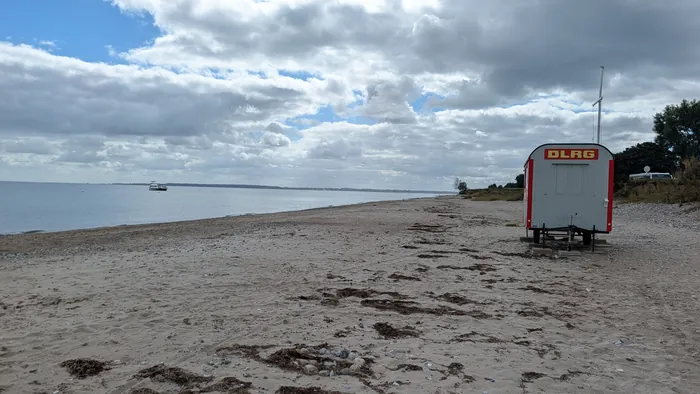 Strand Booknis unter Wolken, rechts der rot-weiße Wagen der DLRG auf dem Sand, davor Tang in Streifen, links das ruhige Wasser der Eckernförder Bucht mit einem Boot