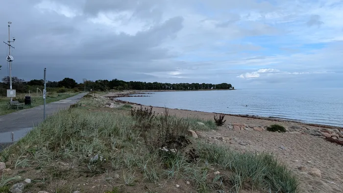 Bucht bei Bülk unter grauen Wolken, links ein Asphaltweg mit Bank und ein Mast mit Messgeräten, im Vordergrund Strandhafer auf der Düne, dahinter der geschwungene Sandstrand mit Steinen und rotbraunem Tang, rechts die offene Ostsee