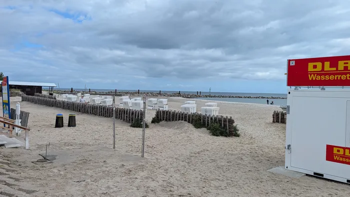 Damp Hundestrand mit weißen Strandkörben hinter niedrigen Faschinenzäunen, rechts der rote Container der DLRG-Wasserrettung, hinten die Steinmolen im Wasser