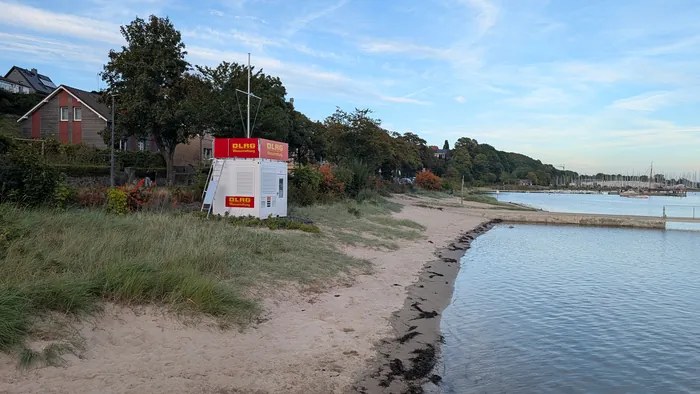Kleiner Sandstrand in Eckernförde Borby, links der weiße Container der DLRG-Wasserrettung zwischen Bäumen, rechts das ruhige Wasser und dahinter die Masten im Yachthafen