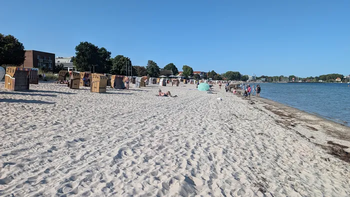 Stadtstrand von Eckernförde mit Reihen bunter Strandkörbe im hellen Sand, dahinter die Stadt mit dem Kirchturm, rechts die Masten im Hafen