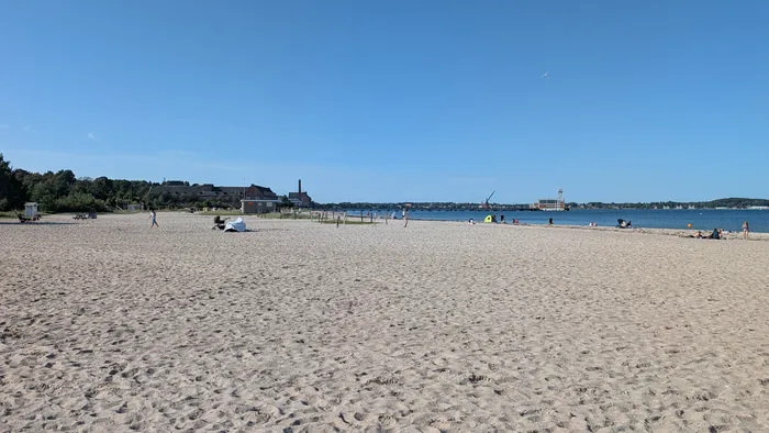 Weiter heller Sandstrand am Eckernförder Südstrand, im Hintergrund die Hafenkante mit Kran und Turm, rechts die Bucht
