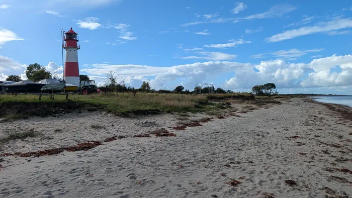 Sandstrand bei Falshöft unter blauem Himmel mit Quellwolken, links der rot-weiße Leuchtturm, davor Boote auf Trailern und ein Traktor, rechts die ruhige Ostsee