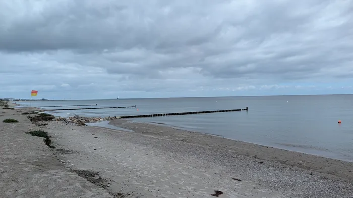 Fischleger Strand bei grauem Himmel, zwei Buhnen aus Holzpfählen ragen ins flache Wasser, links eine rot-gelbe Flagge der Wache und ein Abbruch mit Steinen