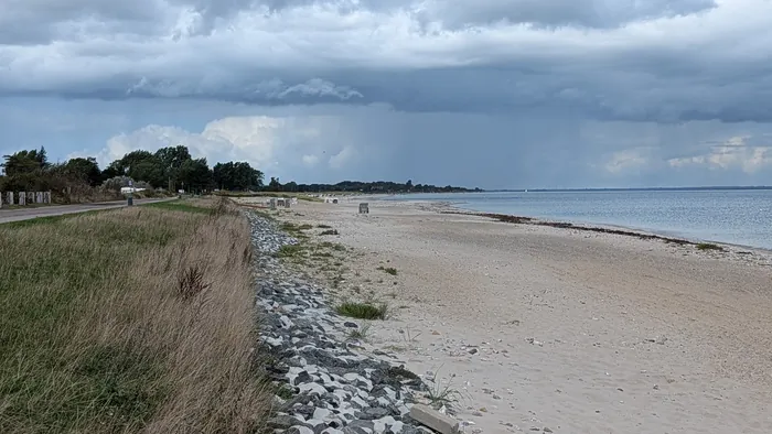 Strand bei Hasselberg unter einer dunklen Regenwolke, links der begrünte Deich mit dem Weg darauf, am Deichfuß eine Steinschüttung, in der Ferne eine Reihe Strandkörbe, rechts die ruhige Ostsee