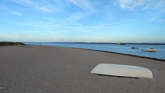 Sandstrand bei Hemmelmark am Abend, vorn ein umgedrehtes weißes Boot auf dem Sand, auf dem Wasser Motorboote und ein Schiff vor Anker, links Dünengras
