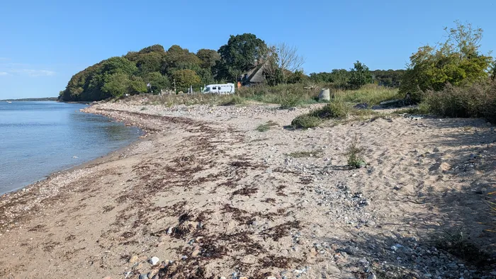 Stein- und Sandstrand bei Kiekut mit dunklem Tang im Spülsaum, oben am Ufer ein Reetdachhaus und ein weißes Wohnmobil, links das flache Wasser der Eckernförder Bucht