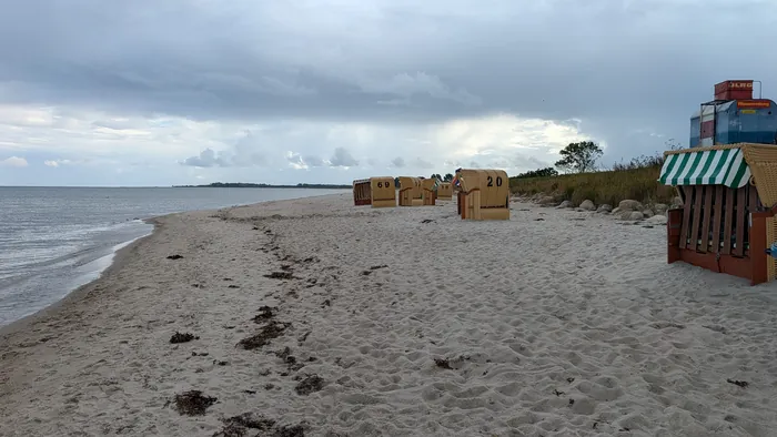 Sandstrand in Kronsgaard an einem bedeckten Tag, in der Mitte eine Reihe nummerierter Strandkörbe, rechts der Turm der DLRG-Wasserrettung, links das flache Wasser der Ostsee