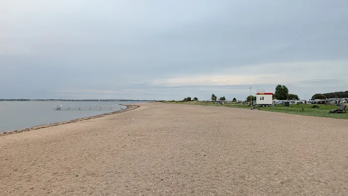 Sehr breiter Sandstrand bei Langballigau unter bedecktem Himmel, links ein Badesteg mit Geländer im flachen Wasser, rechts auf der Wiese ein weißer DLRG-Wagen, dahinter Bäume und die Reihen des Campingplatzes