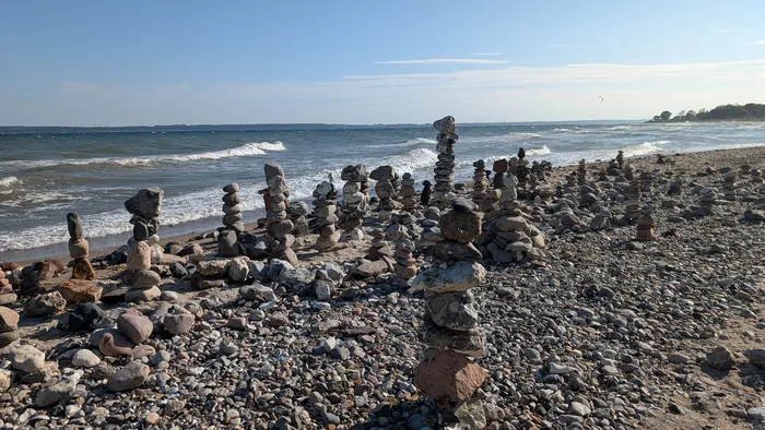 Steinstrand bei Langholz mit vielen aufgetürmten Steinmännchen, dahinter Wellen mit Schaumkronen auf der Eckernförder Bucht, am Horizont ein Kitesurfer