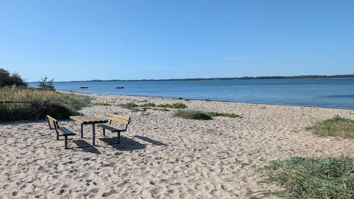 Bank und Tisch aus Holz stehen im Sand am Strand von Lindhöft, dahinter der Spülsaum mit Steinen und zwei kleine Boote auf der Eckernförder Bucht