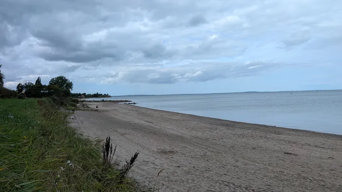 Breiter Sandstrand bei Norgaardholz unter bedecktem Himmel, links eine Grasböschung mit Bäumen, weiter hinten eine Steinbuhne und ein rotes Haus, auf dem Sand sitzt ein Mensch, rechts die Flensburger Förde