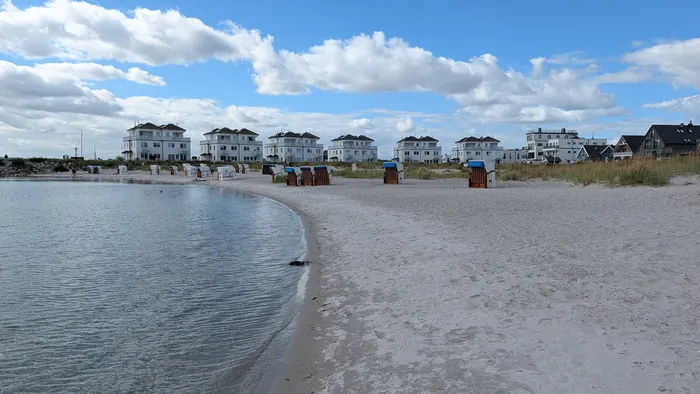 Strand Olpenitz in einer geschwungenen Bucht, Strandkörbe vor Dünengras, dahinter die weißen Ferienhäuser des Ostseeresorts