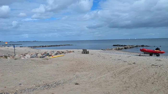 Strand Schönhagen mit Steinbuhnen im flachen Wasser, rechts das rote Rettungsboot der DLRG auf einem Hänger, links die Wachflagge, am Horizont Segelboote und die Häuser von Damp