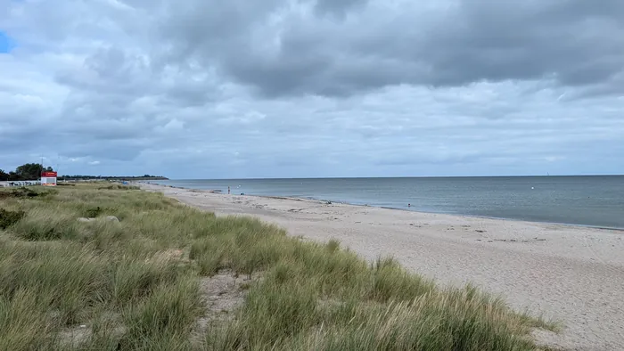 Schubystrand unter Wolken, vorn Dünengras, dahinter der lange helle Sandstrand mit wenigen Badenden, links in der Ferne der Turm der DLRG, rechts die ruhige Ostsee