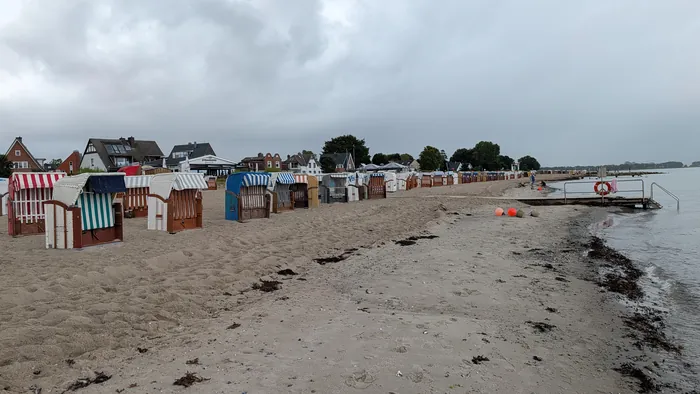 Strand von Strande an einem bedeckten Tag, eine lange Reihe bunt gestreifter Strandkörbe steht im Sand, dahinter die Häuser und ein Restaurant am Ufer, rechts ein Badesteg mit Rettungsring und Geländer im flachen Wasser