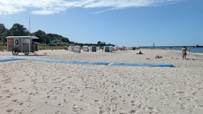 Sandstrand in Surendorf mit dem hölzernen DLRG-Wagen links, quer über den Strand eine blaue Matte als Zugang zum Wasser, dahinter eine Reihe Strandkörbe und rechts die Seebrücke