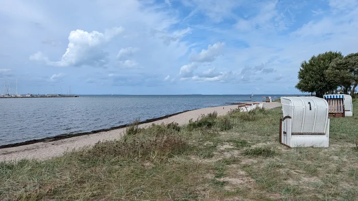 Strand bei Wackerballig unter blauem Himmel mit Quellwolken, rechts weiße Strandkörbe im Strandhafer, links die Mole mit den Masten der Segelboote, auf dem Wasser ein Windsurfer