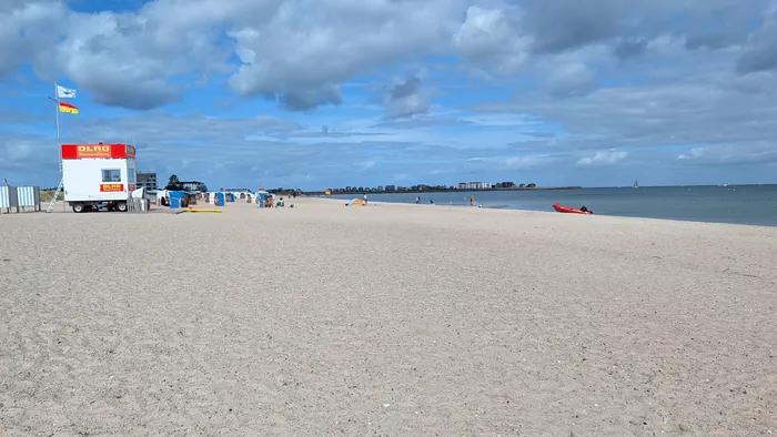 Weidefelder Strand mit breitem Sand, links der Wagen der DLRG-Wasserrettung mit Flaggen und Strandkörbe, rechts das Wasser mit einem roten Rettungsboot, am Horizont die Häuser von Damp