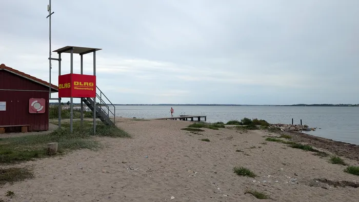 Badestrand bei Westerholz unter bedecktem Himmel, links ein rotes Holzhaus mit Rettungsring und ein roter DLRG-Turm, dahinter ein Holzsteg ins Wasser, von dem eine Person zurück an Land kommt